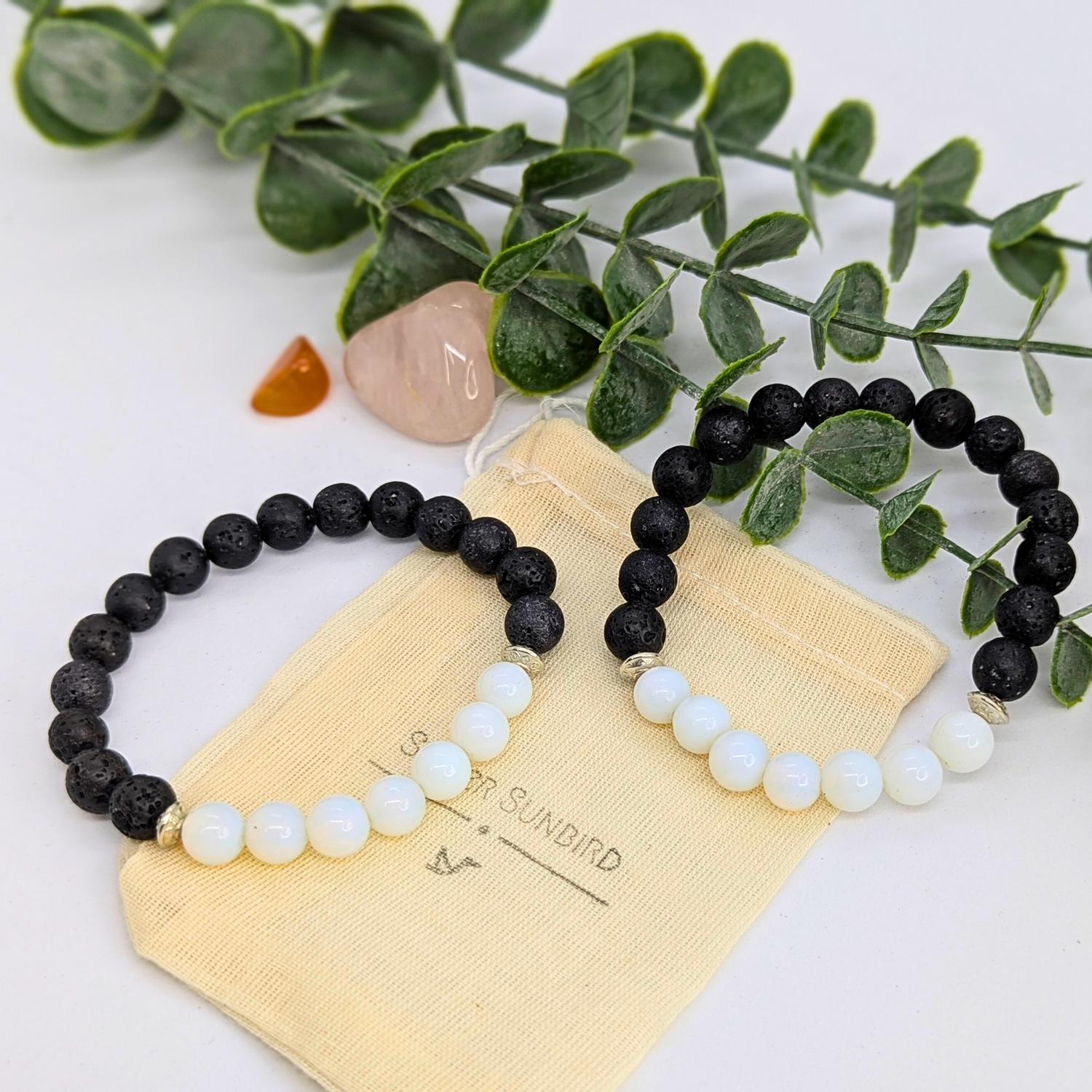 Two Opal and Lava Stone Bracelets by Silver Sunbird rest on a cream pouch labeled “Silver Sunbird,” with a branch of green leaves and two polished stones in the background.