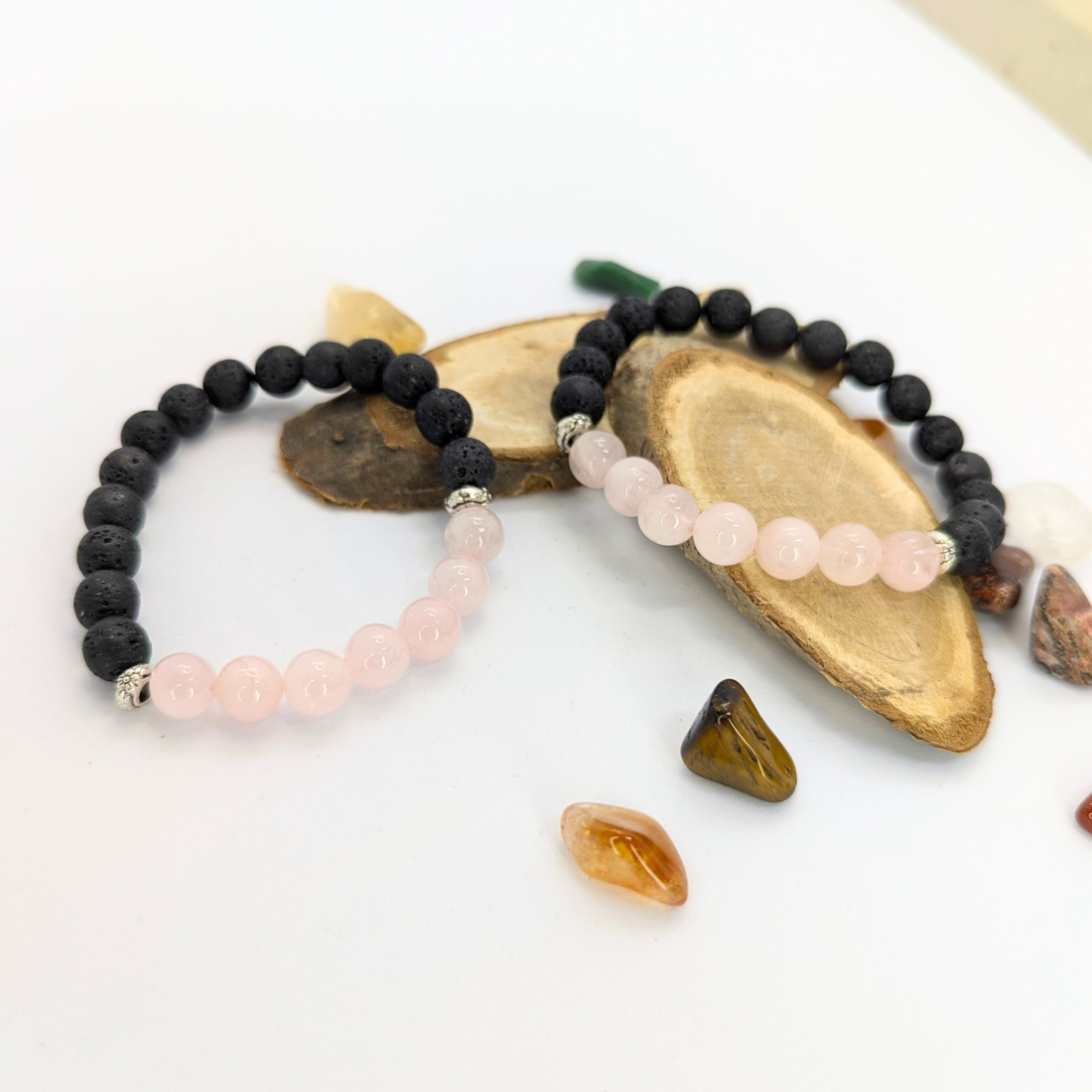 The Rose Quartz and Lava Stone Bracelets by Silver Sunbird elegantly rest on wooden slices. Around them, an assortment of colorful gemstones in various shapes is artfully scattered on a white surface.