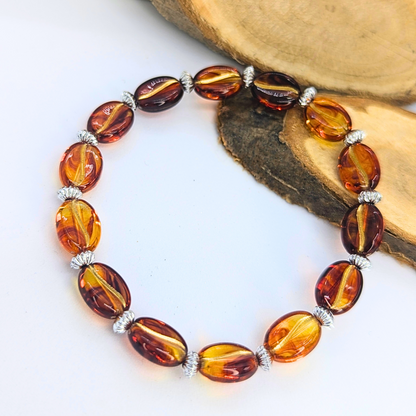 The Silver Sunbird Coffee Bean Bracelets, crafted with polished amber beads and silver spacers, are showcased on a white surface. Wooden slabs peek into view, bringing in a natural touch reminiscent of the exquisite bracelets cherished by coffee enthusiasts.