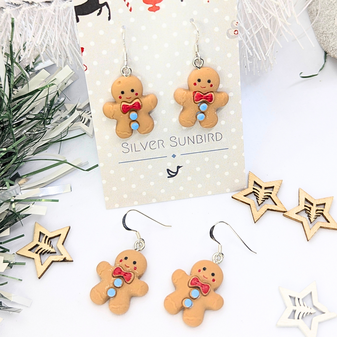 Displayed on a white card labeled "Silver Sunbird," the Cheerful Gingerbread Man Earrings are adorned with red bow ties and blue buttons. The background features festive decor, including white branches and wooden star ornaments.