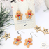 Displayed on a white card labeled "Silver Sunbird," the Cheerful Gingerbread Man Earrings are adorned with red bow ties and blue buttons. The background features festive decor, including white branches and wooden star ornaments.
