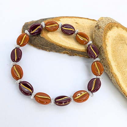 The Silver Sunbird Coffee Bean Bracelets are exquisite with alternating maroon and brown oval beads featuring gold stripes, separated by small silver beads and arranged on light brown wood—perfect for coffee enthusiasts.