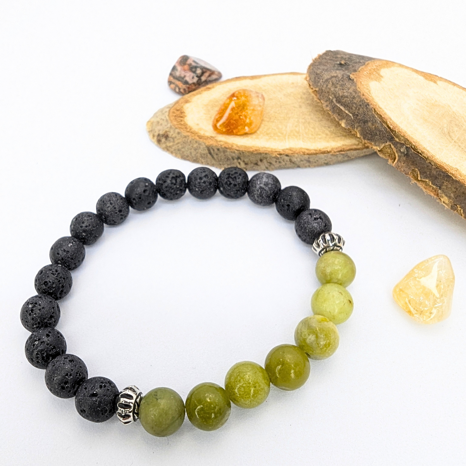 The Silver Sunbird Peridot Bracelet, featuring black lava stones and olive green gems with two silver spacers, exemplifies exquisite gemstone jewelry. Nearby lie three polished stones—amber, brown with orange speckles—resting on wooden slices against a white background.