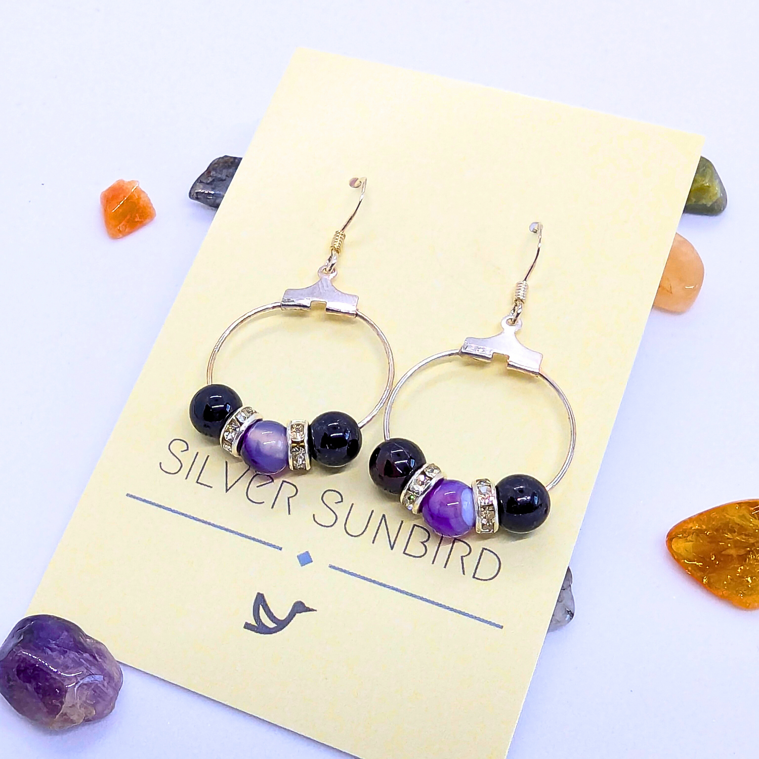 A pair of Amethyst Statement Large Earrings adorned with purple  gemstones and silver rhinestone spacers is displayed on a card labelled &quot;Silver Sunbird.&quot; The card is set against a white background, scattered with small natural gemstones in orange, green, and purple hues.