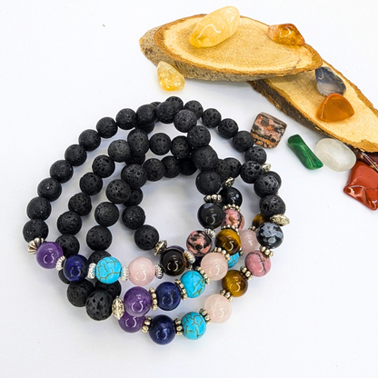 The Chakra Bracelet by Silver Sunbird, crafted with black lava stones and semi-precious gemstones, is displayed coiled on a white surface beside wood slices and colorful polished crystal stones.