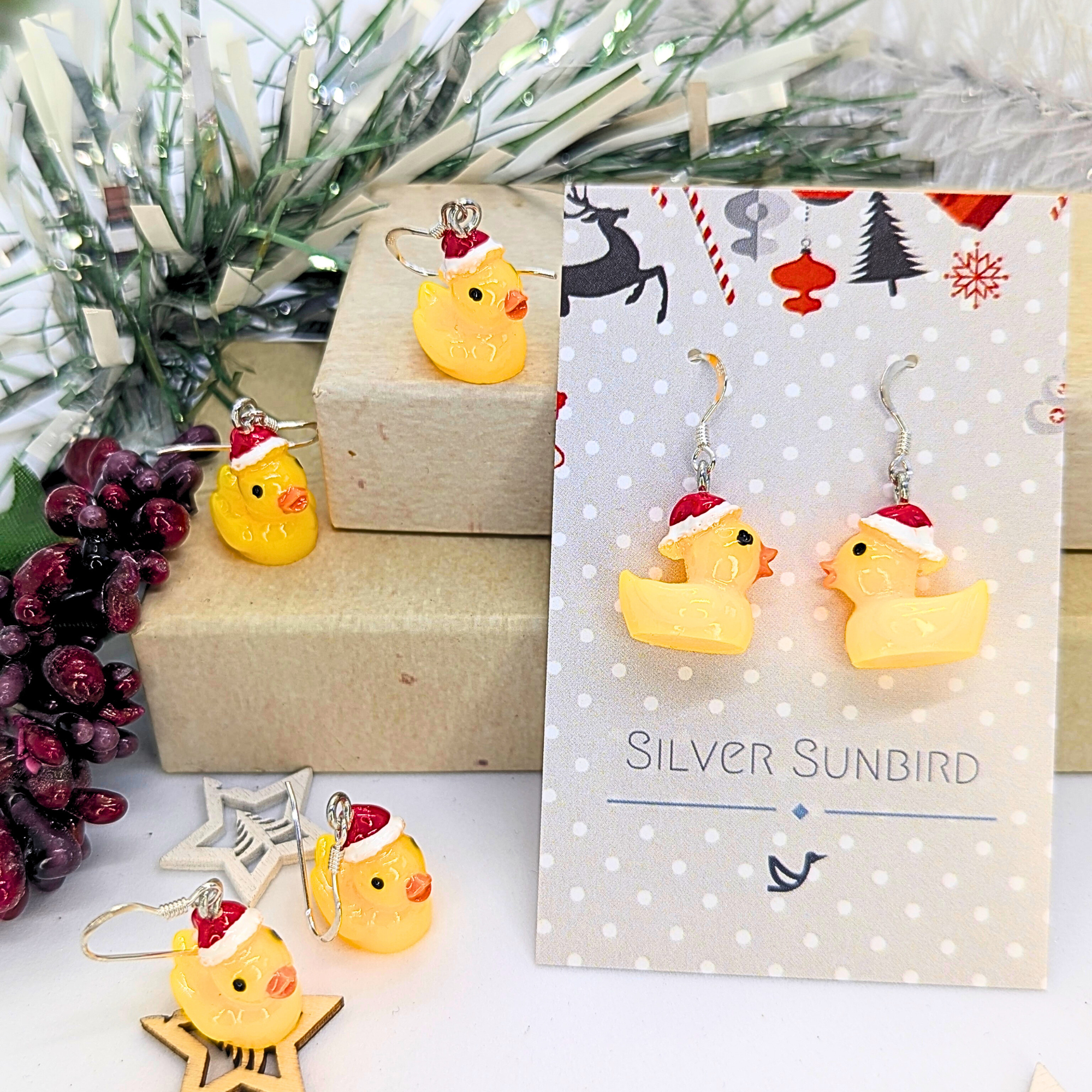 A close-up showcases the Silver Sunbird Festive Duck Earrings, featuring handcrafted yellow ducks adorned with Santa hats, elegantly hanging from sterling silver hooks. These earrings are presented on a cheerful card embellished with a pine branch and red berries in the background, making them an ideal holiday statement piece.