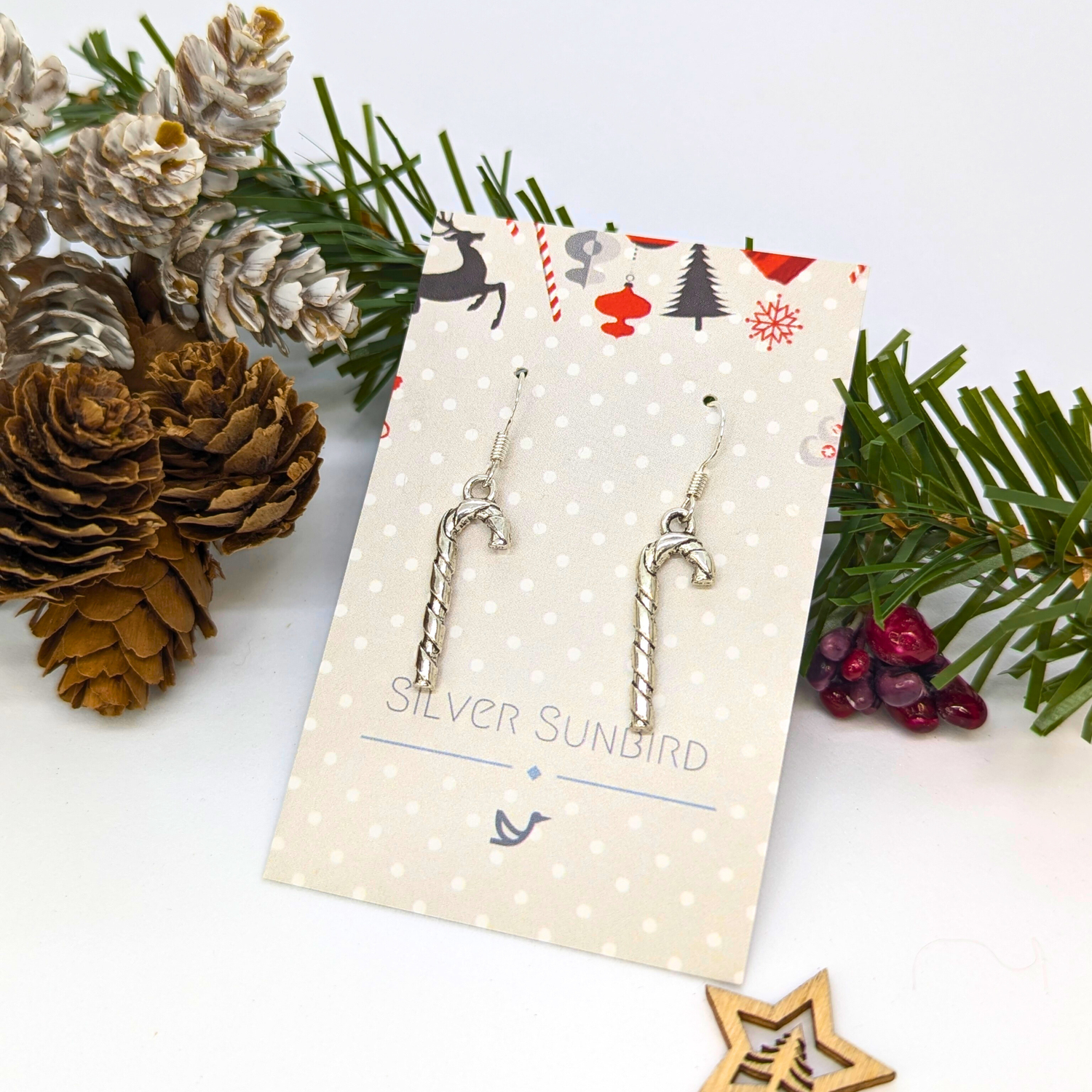 Classic Candy Cane Earrings by Silver Sunbird, crafted from sterling silver, are displayed on a polka dot card with festive Christmas décor—pinecones and evergreens—in the background.