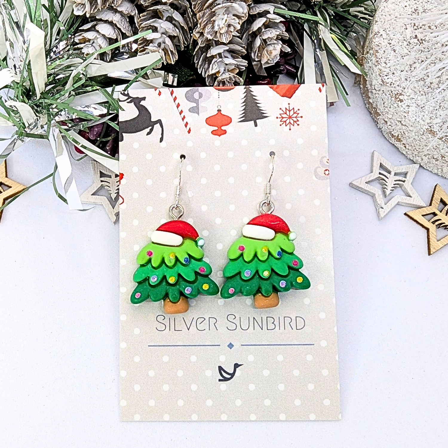 The Silver Sunbird Christmas Tree Earrings are adorned with red Santa hats and elegantly mounted on a Silver Sunbird card. The display is enhanced by a background of pine cones, greenery, and star decorations, creating the perfect holiday accessory.