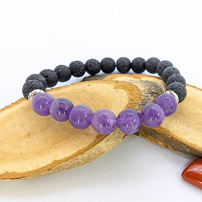 A stretch bracelet made with black lava stones and polished purple amethyst beads is displayed on a wooden slice. The amethyst beads are glossy and vibrant, contrasting with the textured, porous appearance of the lava stones. A small orange polished stone is visible in the background, adding a touch of colour to the composition.