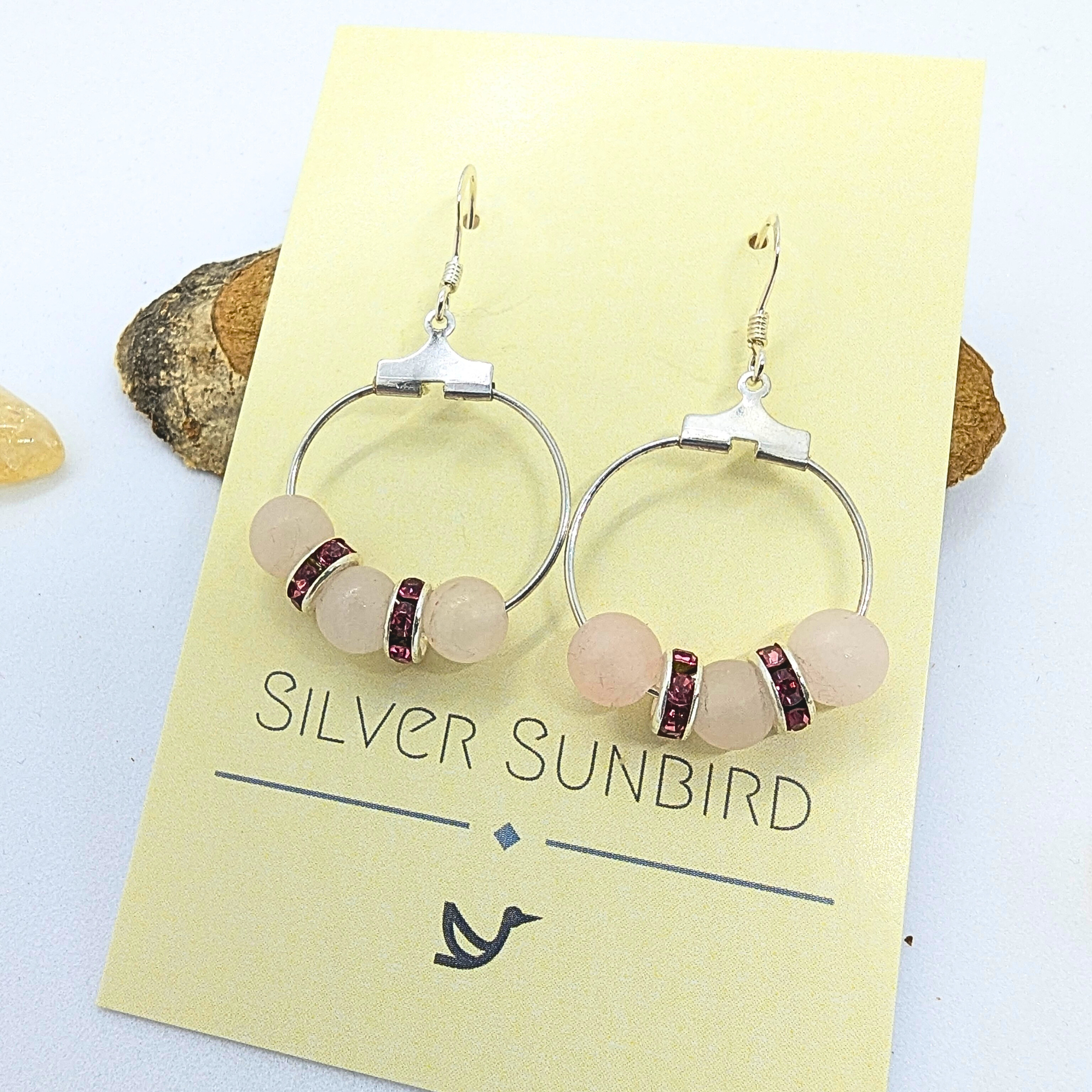 A pair of Rose Quartz Statement Earrings from Silver Sunbird displayed on a yellow card. Each earring features a silver hoop with three light pink rose quartz gemstones and two smaller maroon rhinestone spacer beads. The card shows the brand name &quot;Silver Sunbird&quot; and a simplistic bird logo at the bottom.