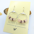 A pair of Rose Quartz Statement Earrings from Silver Sunbird displayed on a yellow card. Each earring features a silver hoop with three light pink rose quartz gemstones and two smaller maroon rhinestone spacer beads. The card shows the brand name "Silver Sunbird" and a simplistic bird logo at the bottom.