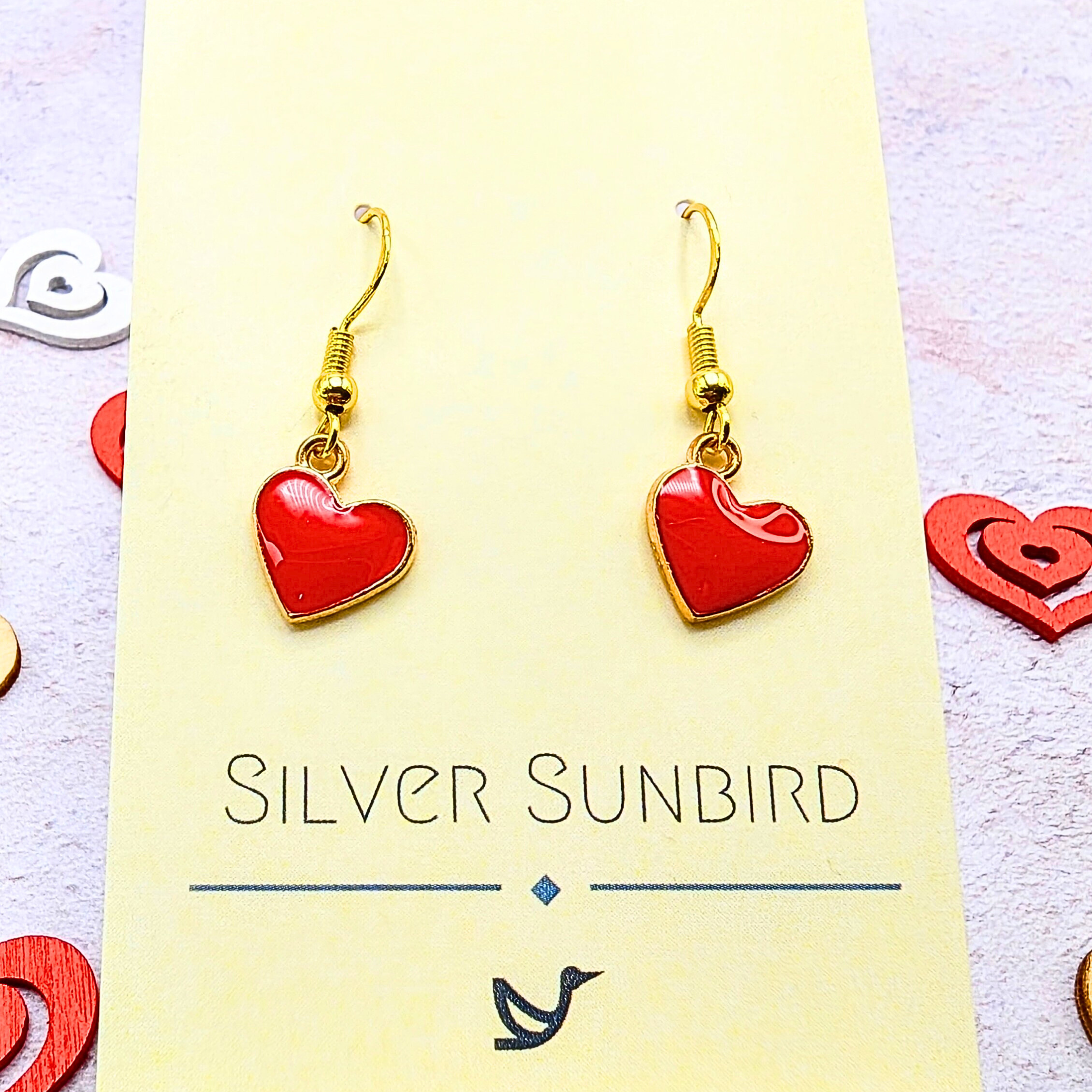 The Alluring Red Heart Earrings by Silver Sunbird dangle elegantly on a cream card and are adorned with heart-shaped pendants. Surrounded by scattered heart decorations, these hypoallergenic earrings add romantic charm to any setting.