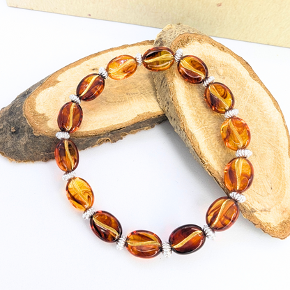 The Silver Sunbird Coffee Bean Bracelet, with its alternating amber and silver beads, forms a circle on two overlapping wooden pieces against a plain background. The glossy, oval-shaped amber beads captivate with their warm glow.