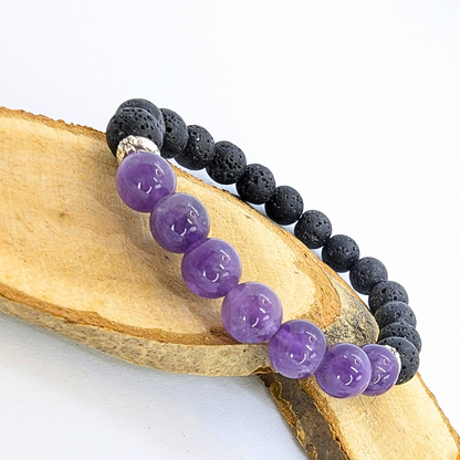 A stretch bracelet made with black lava stones and polished purple amethyst beads is displayed on a wooden slice. The amethyst beads are glossy and vibrant, contrasting with the textured, porous appearance of the lava stones.