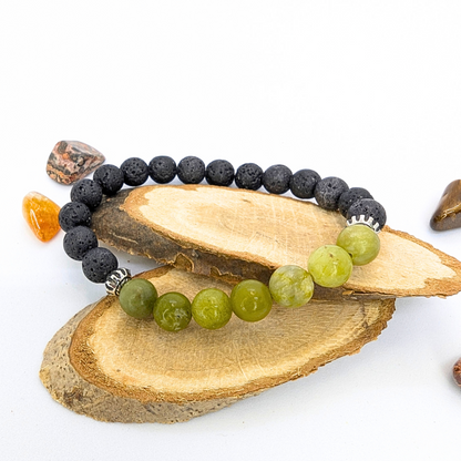 The Silver Sunbird Peridot Bracelet beautifully transitions from black lava to green peridot stones, displayed on wooden slices with small polished brown and orange stones set against a white backdrop highlighting this stunning gemstone jewelry ensemble.