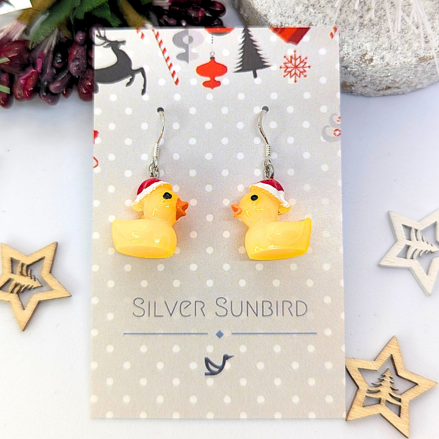 Earrings featuring yellow rubber duck charms wearing red Santa hats are set against a festive backdrop, adorned with wooden star decorations. The text reads &quot;Silver Sunbird.&quot; Explore our Silver Sunbird Festive Duck Earrings for a distinctly unique holiday accessory.