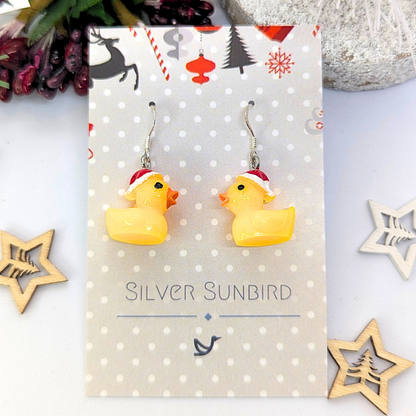 Earrings featuring yellow rubber duck charms wearing red Santa hats are set against a festive backdrop, adorned with wooden star decorations. The text reads &quot;Silver Sunbird.&quot; Explore our Silver Sunbird Festive Duck Earrings for a distinctly unique holiday accessory.