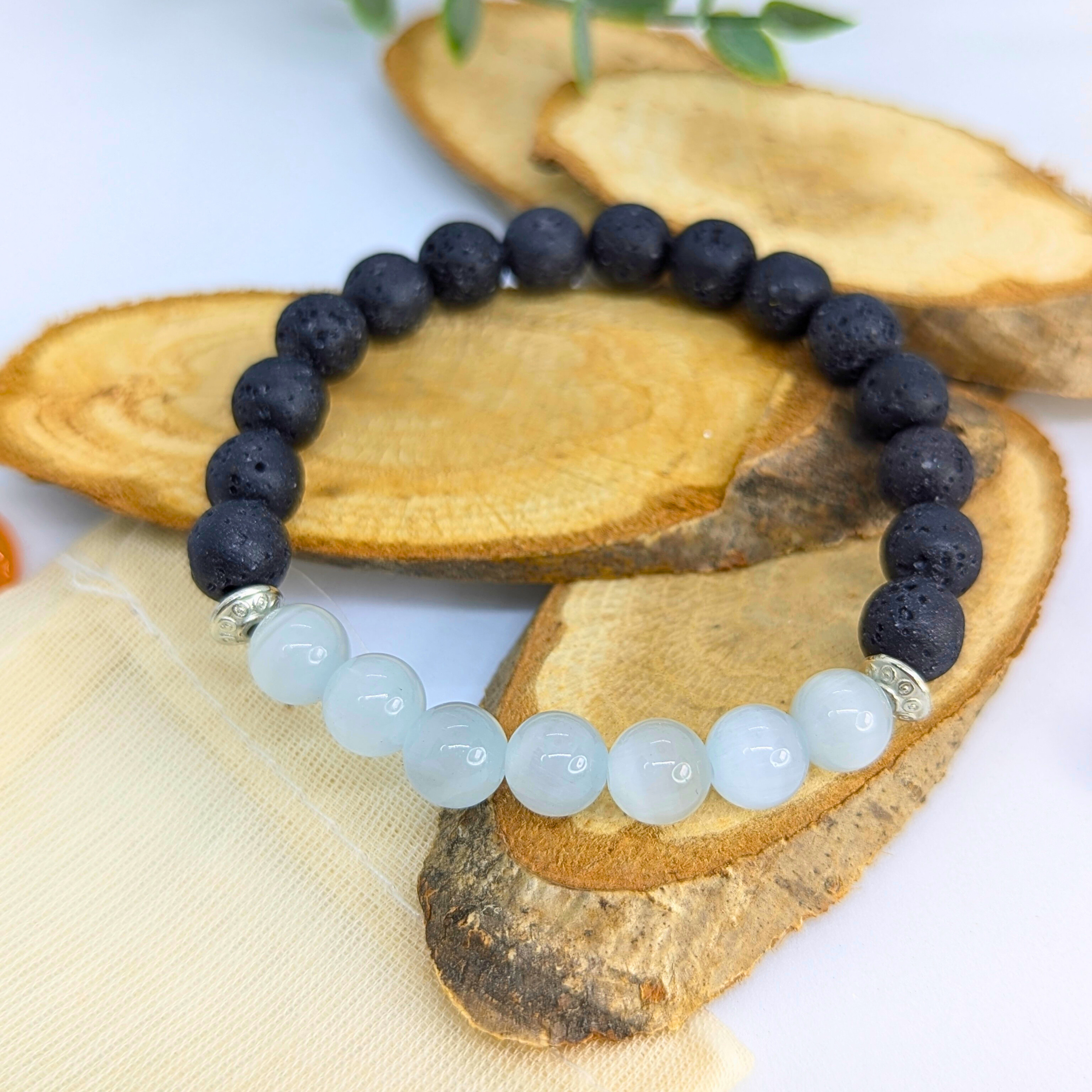 The Silver Sunbird Moonstone and Lava Bracelets, featuring black lava stones, blue-green beads, and silver accents, are displayed on layered wooden slices with a small beige pouch in the background.