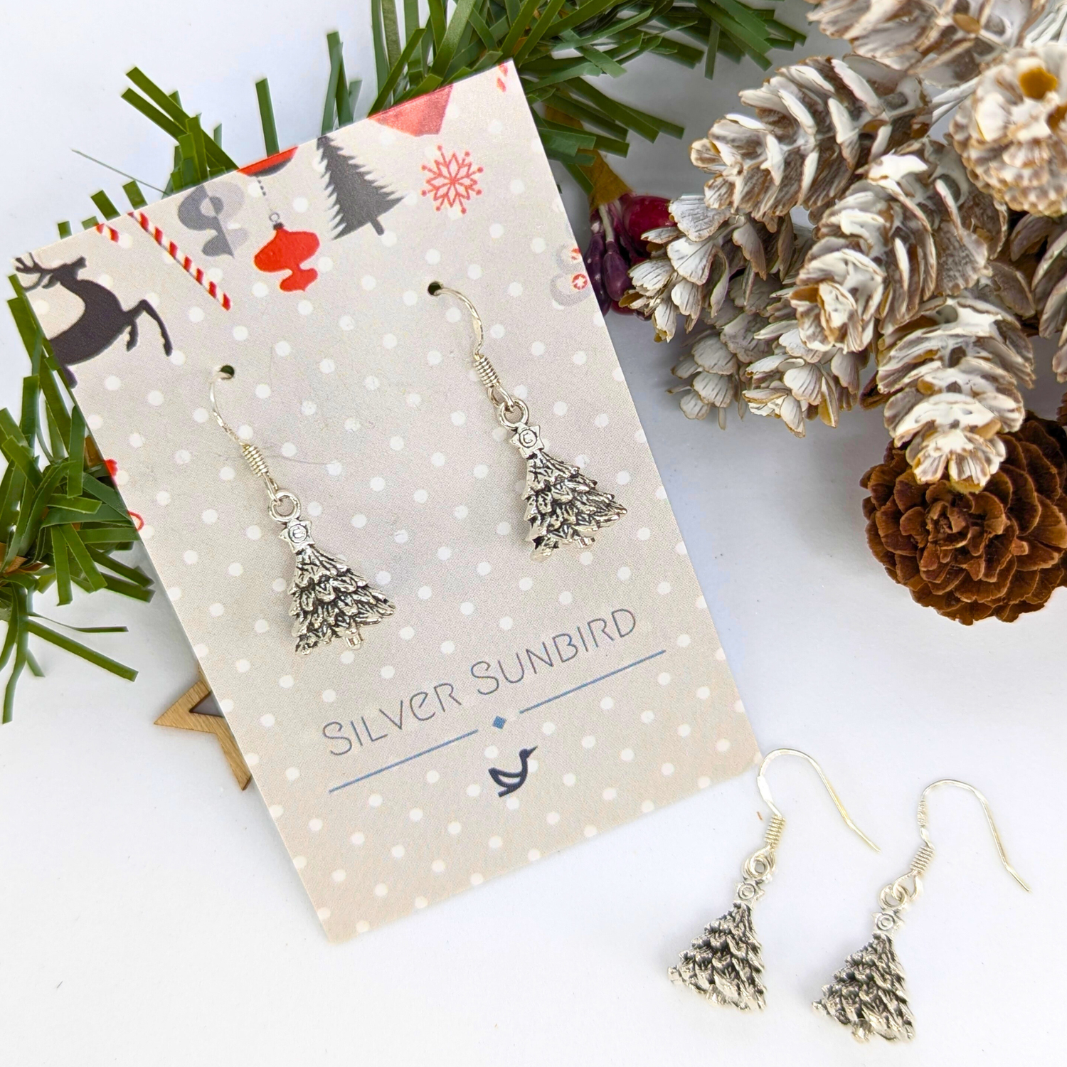 The Silver Sunbird 3D Christmas Tree Earrings are displayed on a festive card beside pine branches, pinecones, and another sterling silver pair, all arranged on a white surface.