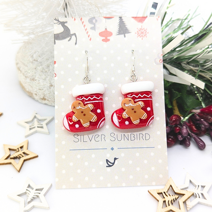Displayed on an elegant &quot;Silver Sunbird&quot; decorative card, the Gingerbread Men Stocking Earrings feature a festive design with white trim and gingerbread figures. Surrounded by holiday-themed decorations and foliage, they are the perfect holiday accessory.