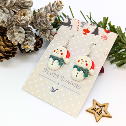 Bright Snowmen Earrings by Silver Sunbird, featuring scarves and Santa hats, are displayed on a polka dot card. Christmas accents like pinecones, frosted greenery, and a wooden star ornament create a festive scene.