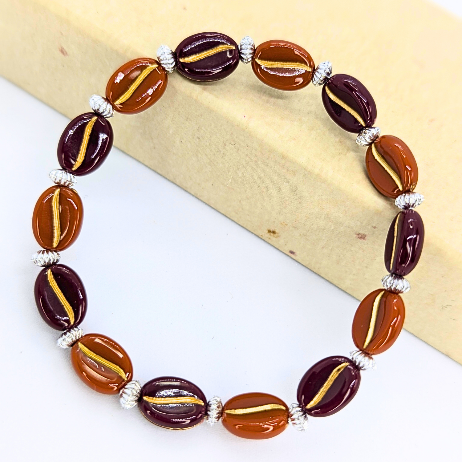 The Coffee Bean Bracelets by Silver Sunbird feature exquisite alternating dark purple and brown beads with gold accents, reminiscent of coffee beans. Elegantly crafted with silver spacer beads, they rest beautifully on a light surface, perfect for coffee enthusiasts.