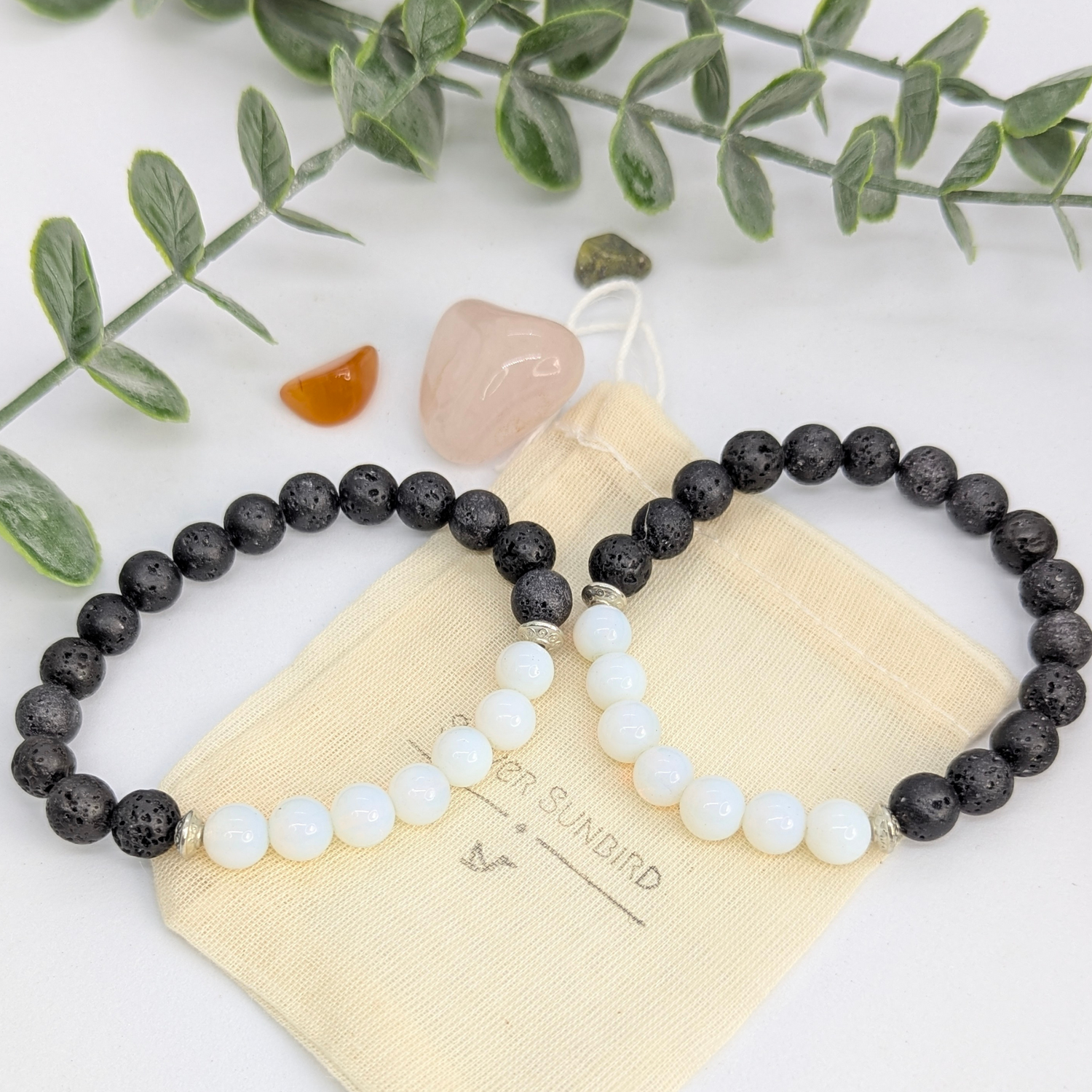 Two Silver Sunbird Opal and Lava Stone Bracelets are displayed on a beige pouch, with polished stones and green leafy stems set against a white background. One bracelet features black and white beads; the other is mostly black.