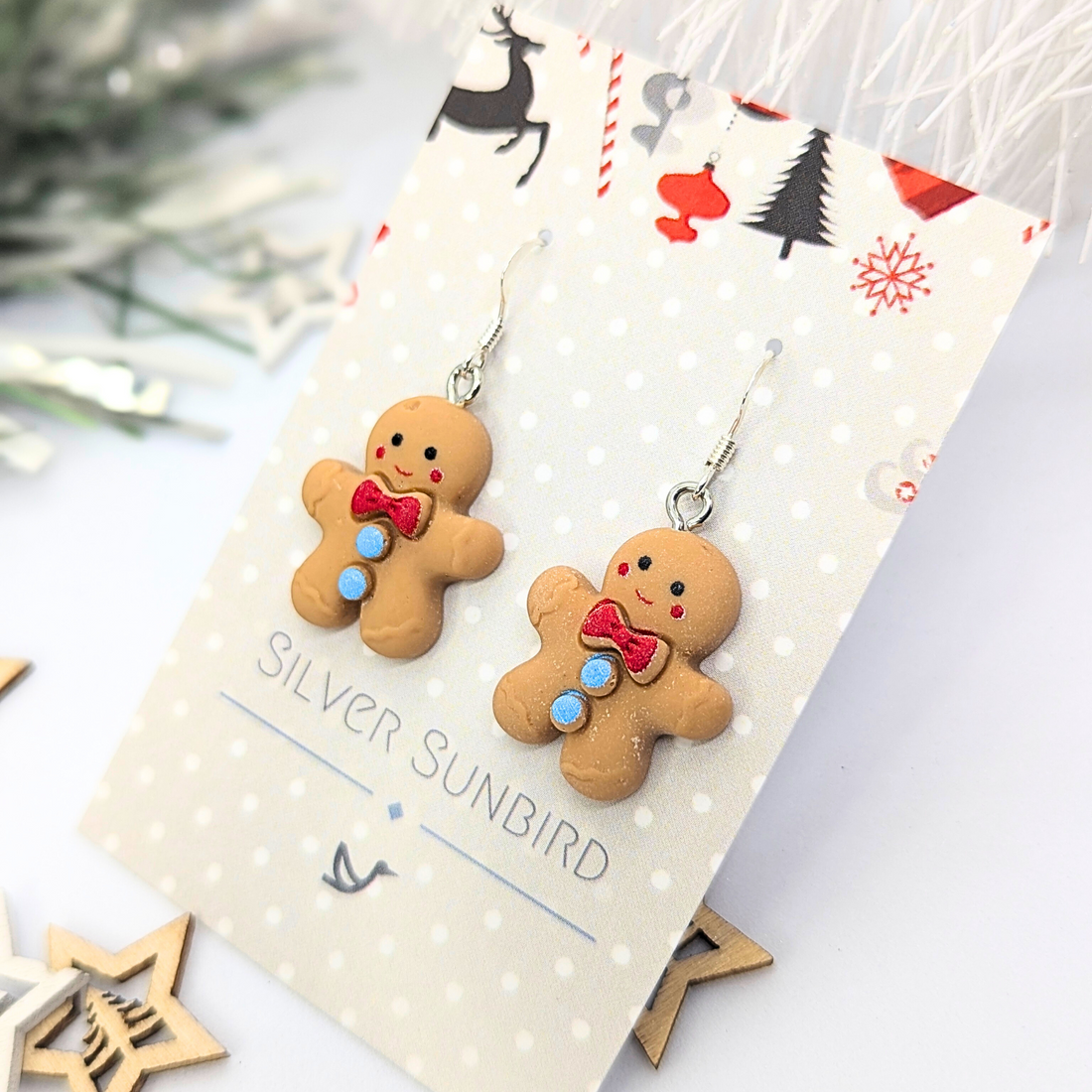 Cheerful Gingerbread Man Earrings featuring red bows and blue buttons, presented on a Christmas-themed display card labeled &quot;Silver Sunbird,&quot; set against a festive background adorned with stars and reindeer.