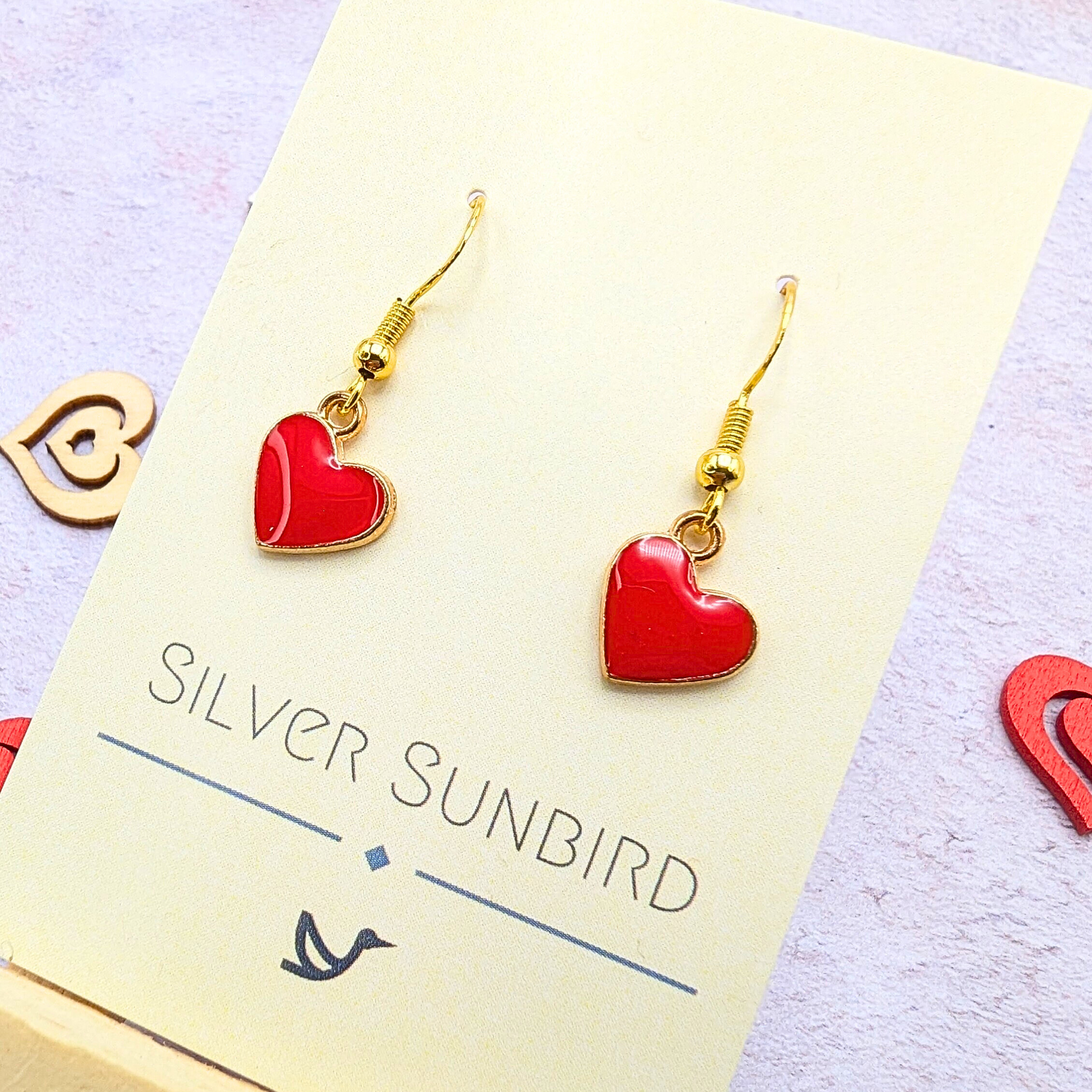 A pair of Alluring Red Heart Earrings by Silver Sunbird, hypoallergenic gold hooks with red heart pendants, displayed on a card. Decorative heart shapes in the background.