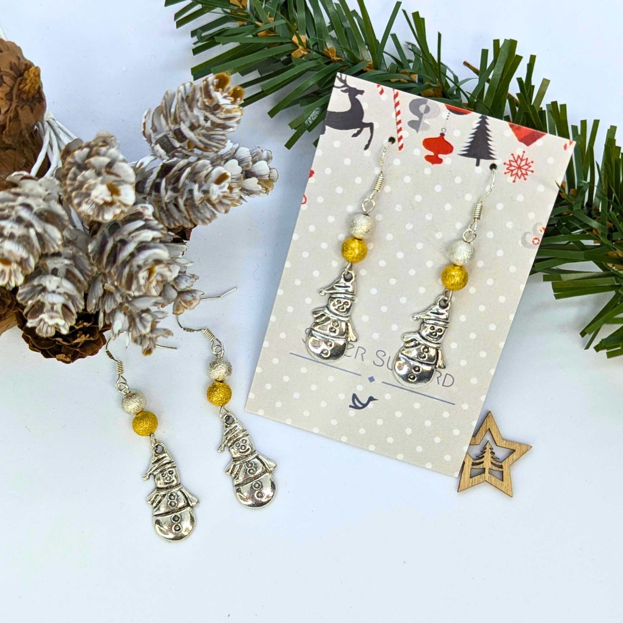 The Very Merry Snowmen Earrings by Silver Sunbird, featuring gold and pearl beads, are shown on a decorative card amid pine cones, frosted greenery, and a wooden star ornament on a white surface.
