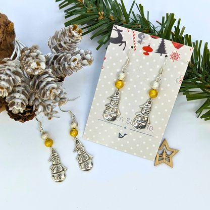 The Very Merry Snowmen Earrings by Silver Sunbird, featuring gold and pearl beads, are shown on a decorative card amid pine cones, frosted greenery, and a wooden star ornament on a white surface.