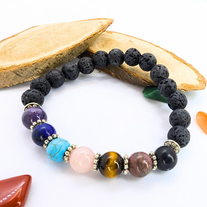 The Chakra Bracelet by Silver Sunbird features black lava stones and colorful semi-precious gemstones, displayed on a white background with two wooden slabs and assorted polished stones surrounding it.