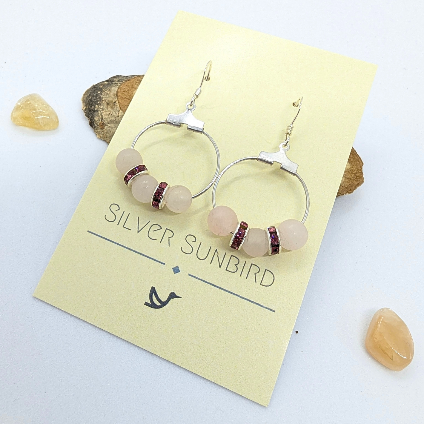 A pair of Rose Quartz Statement Earrings by Silver Sunbird, crafted from sterling silver and featuring a circular design with rose quartz gemstones and small red accents, displayed on a cream-colored card. The hypoallergenic earrings are surrounded by three small, light-brown stones on a white surface.