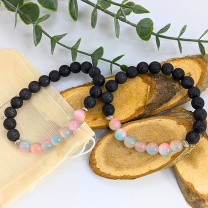 The Silver Sunbird Mermaid-Inspired Lava Stone Bracelet, featuring pastel beads with black lava stones and pink and blue accents, rests on wooden slices. A cloth pouch and green leafy branches complete the serene setting.