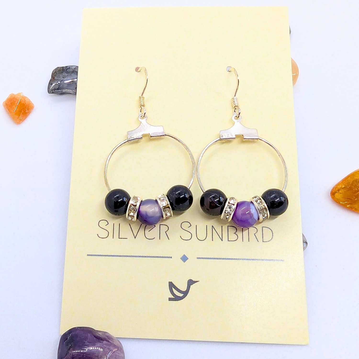 A pair of elegant Amethyst Large Statement  Earrings displayed on a yellow card labelled &quot;Silver Sunbird.&quot; The earrings feature purple amethyst round gemstones, accented with silver rhinestone spacers. The card is adorned with small decorative stones in various colors around its edges.