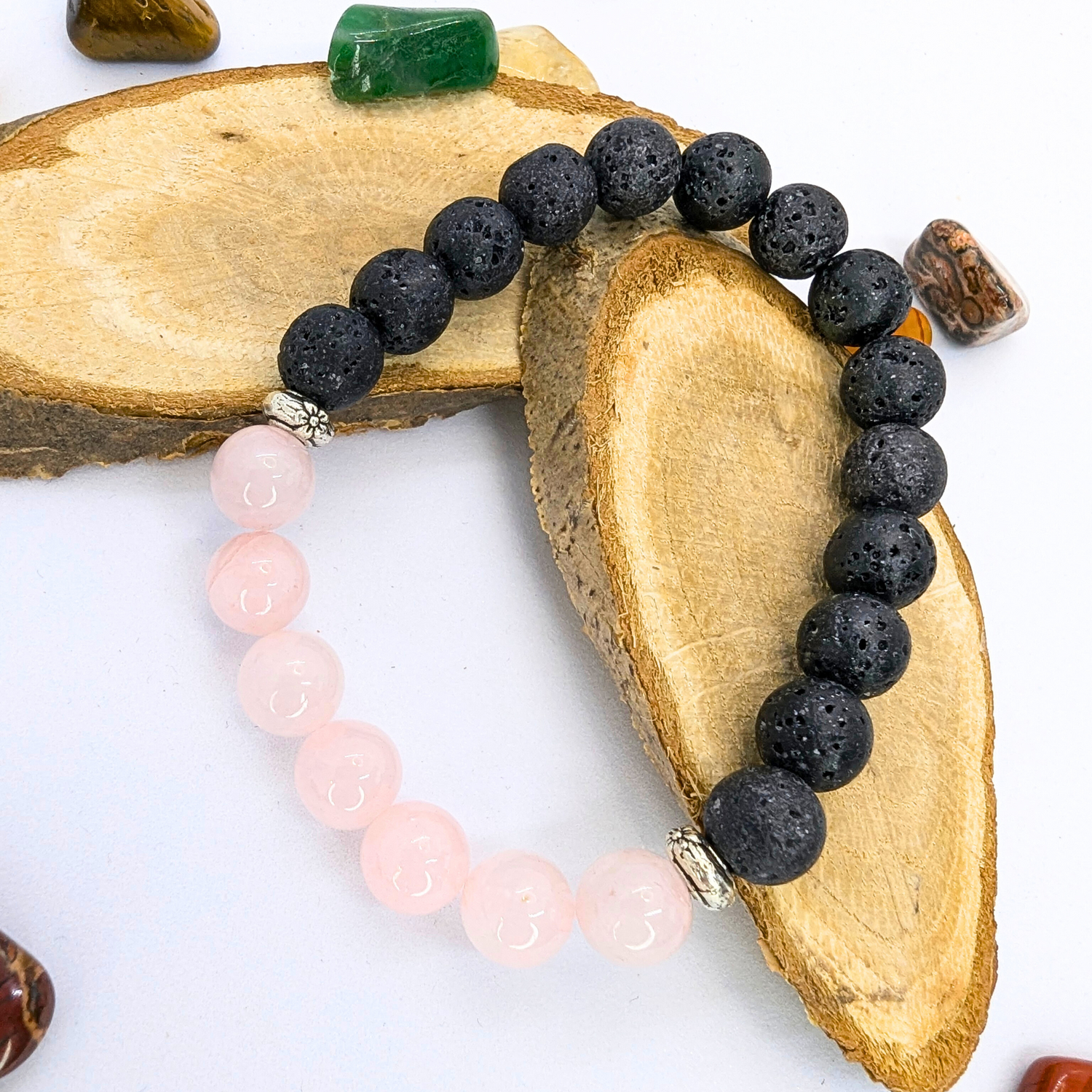 Displayed on wooden slices, the Silver Sunbird Rose Quartz and Lava Stone Bracelets showcase black lava stones and pink rose quartz beads, accented with small silver separators. The presentation includes small polished stones elegantly arranged on a white surface.