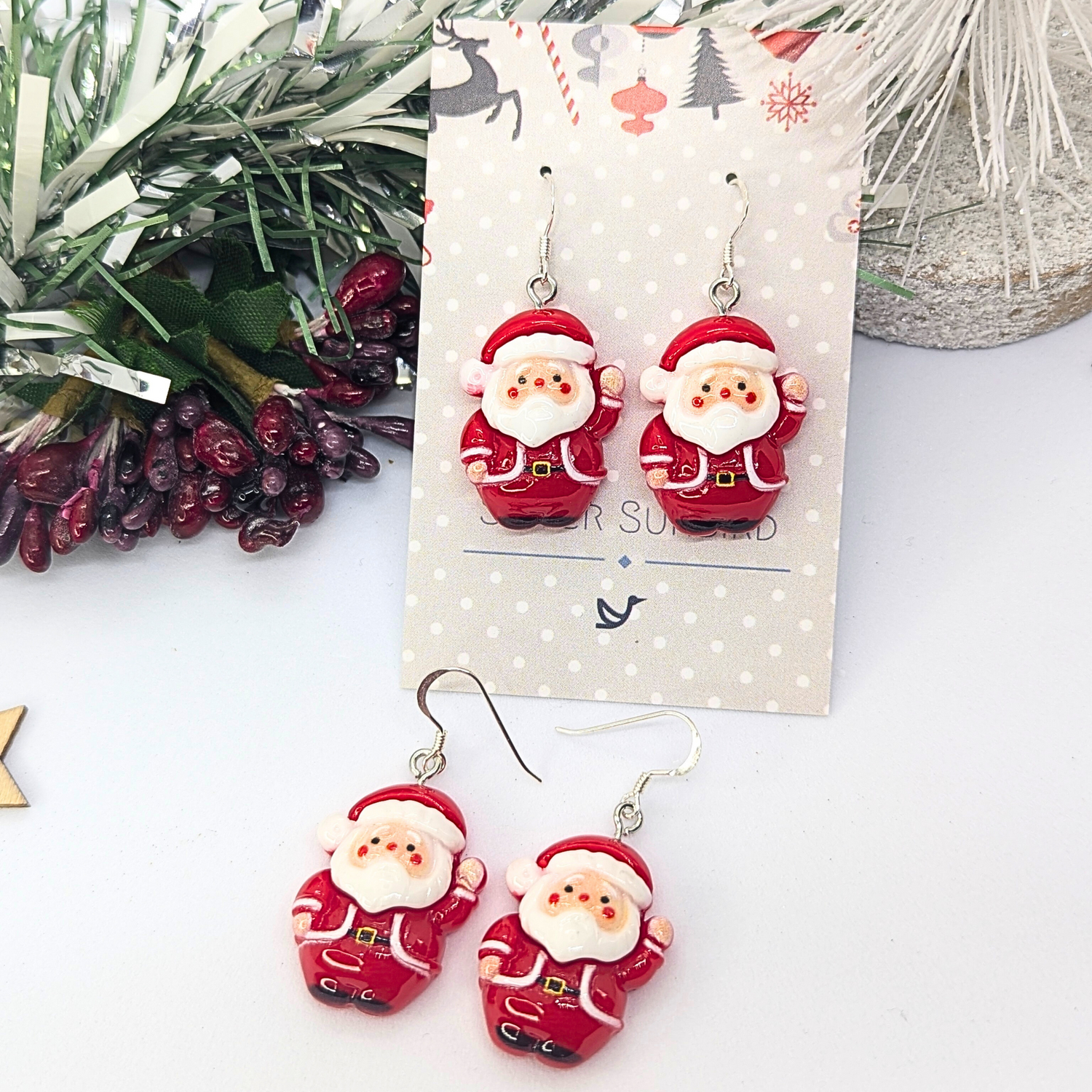 The Jolly Santa Earrings by Silver Sunbird feature a delightful depiction of Santa Claus, each wearing a classic red suit and hat. They are presented on a festive jewelry card that captures the spirit of the season with an arrangement of pine branches and berries, making them ideal for spreading holiday cheer.