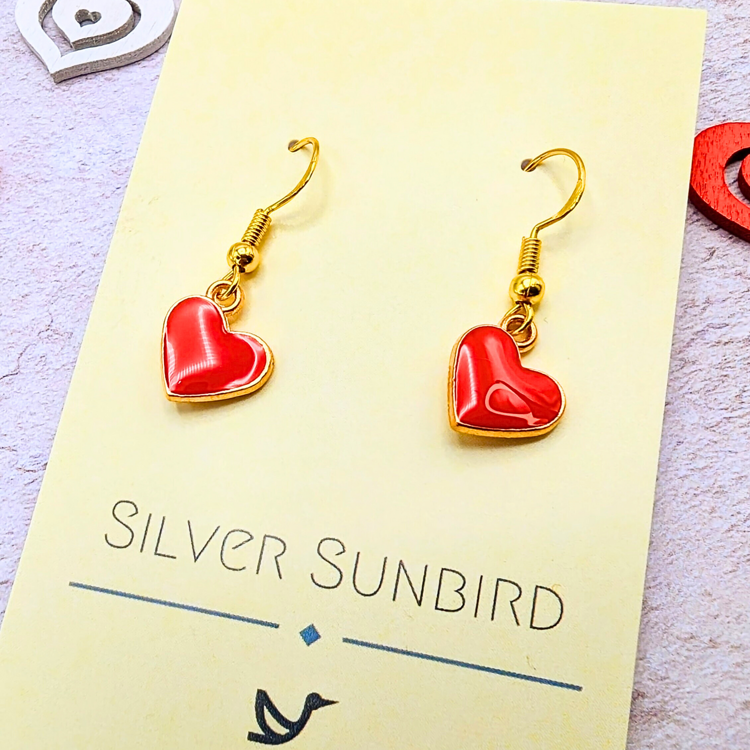 The Alluring Red Heart Earrings by Silver Sunbird feature gold earrings with red heart-shaped pendants, elegantly displayed on a beige &quot;Silver Sunbird&quot; card. Hypoallergenic and perfect for any look, they&