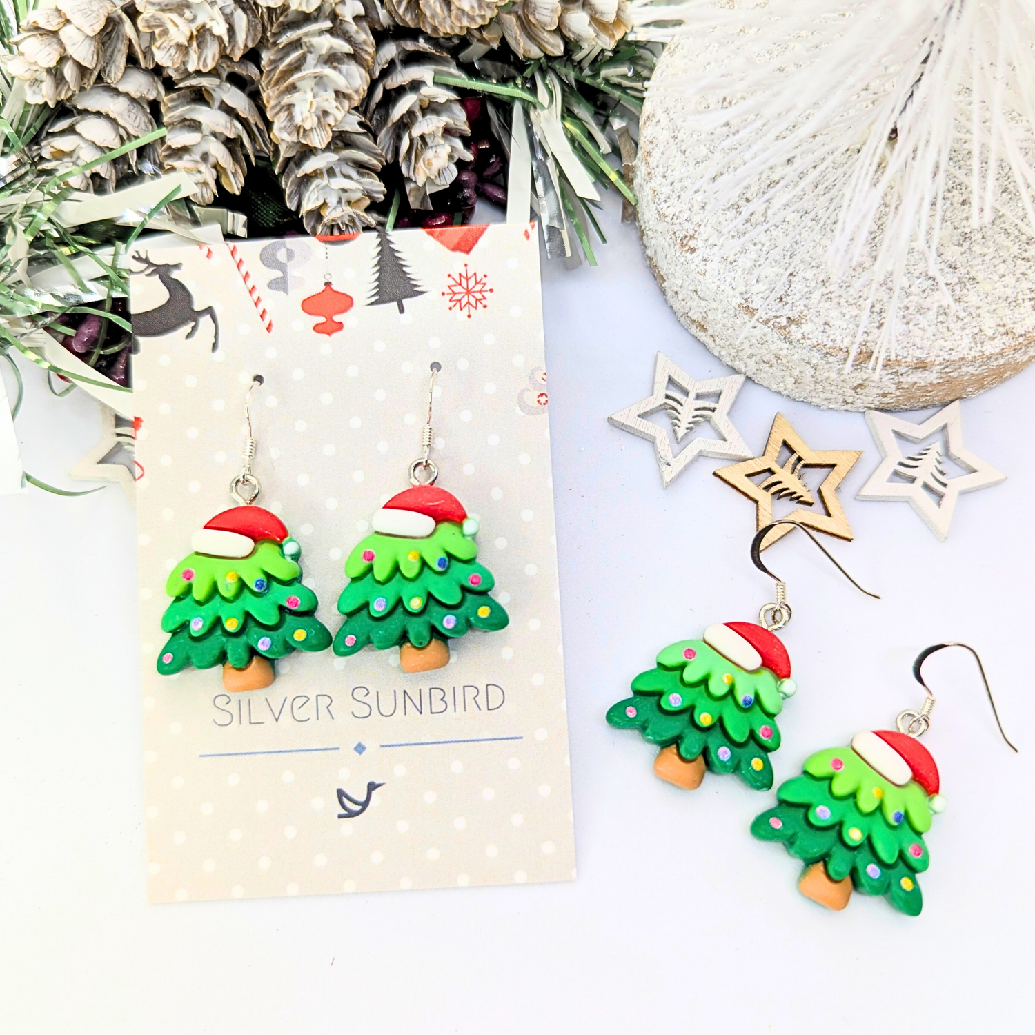 Christmas Tree Earrings by Silver Sunbird, featuring festive designs topped with Santa hats, are beautifully presented on a decorative card. This holiday jewelry is artistically displayed among pine cones, greenery, and wooden star ornaments for a perfect festive touch.