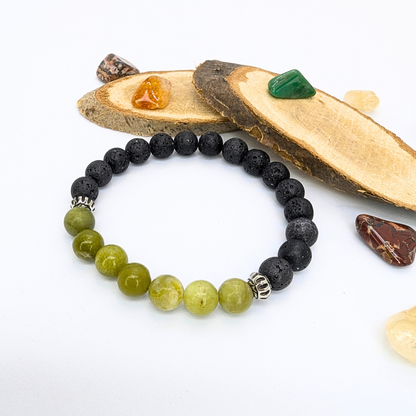 The Silver Sunbird Peridot Bracelet, with its green beads and black lava stones, is beautifully showcased on a white surface amidst wooden slices and colorful stones, highlighting its natural elegance.