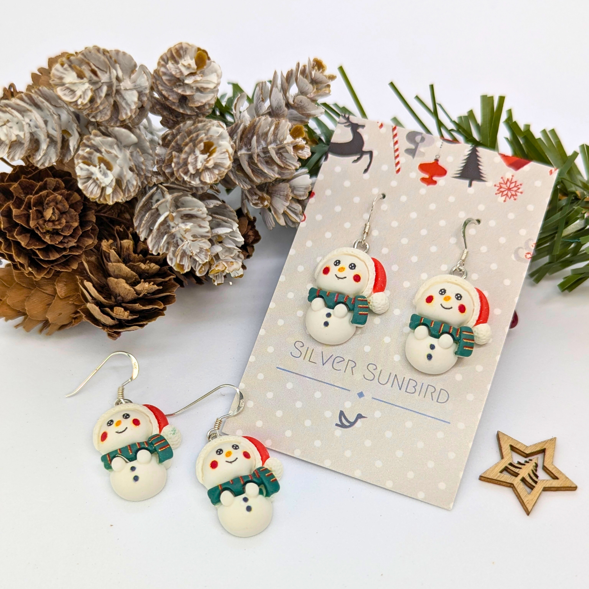 The Bright Snowmen Earrings by Silver Sunbird feature snowman shapes with red hats and green scarves, displayed on a festive branded card with pinecones, pine needles, and a wooden star ornament for a cheerful holiday touch.