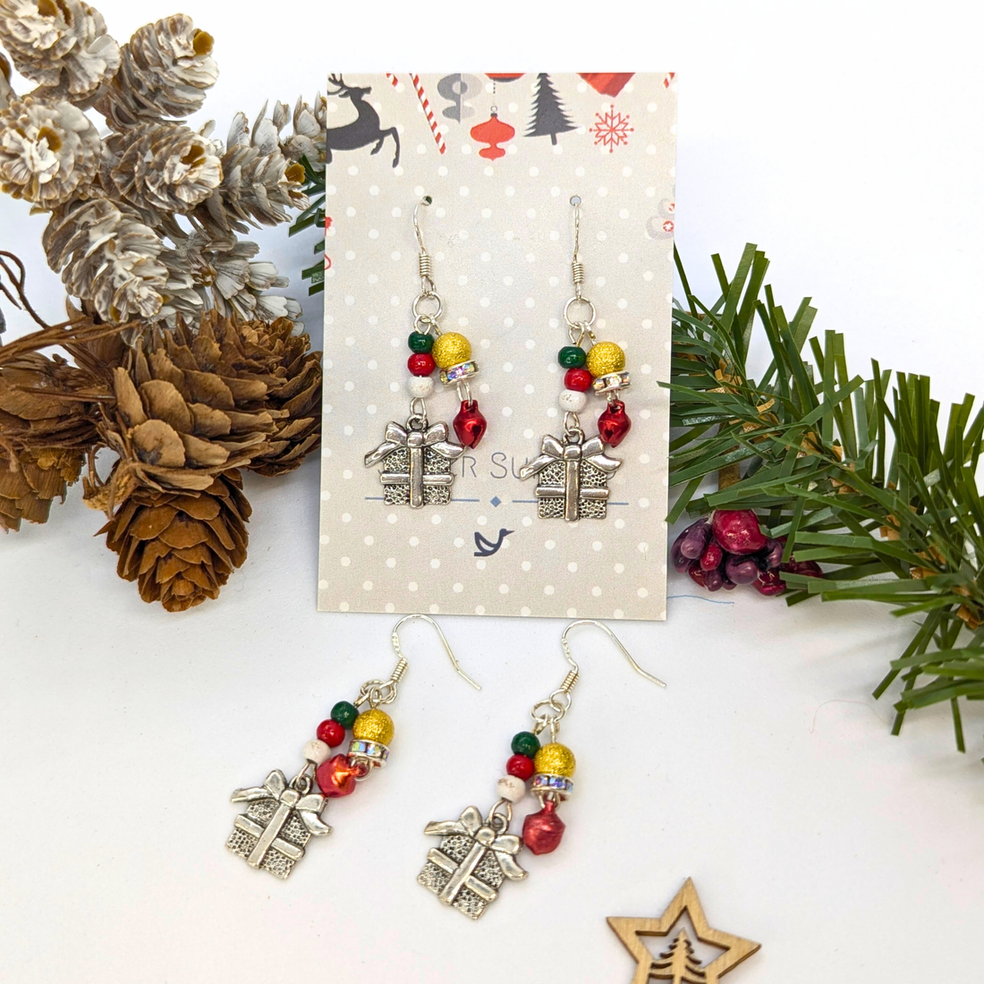Gift Box Dangle Earrings by Silver Sunbird feature sterling silver hooks, silver present charms, and colorful beads. Displayed with pinecones and evergreens, each earring card is adorned with a snowflake pattern.