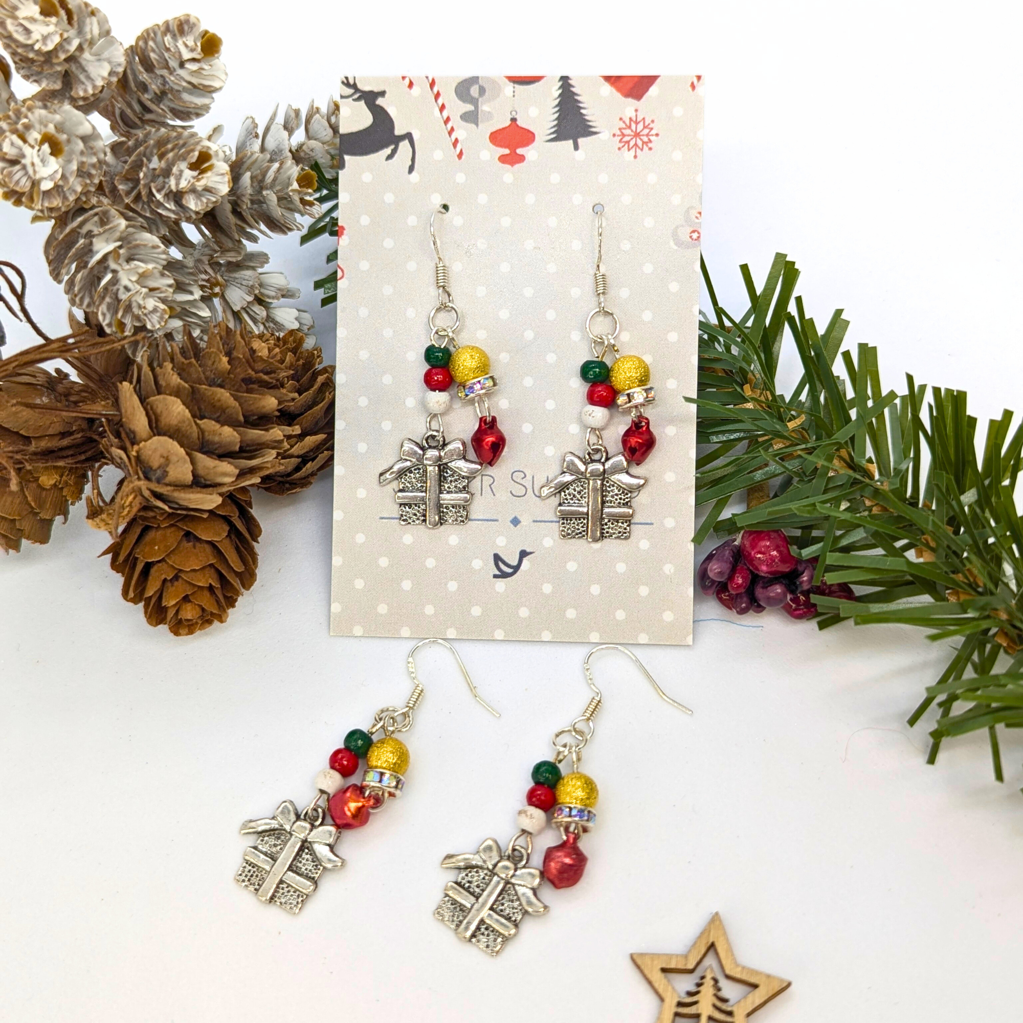 Gift Box Dangle Earrings by Silver Sunbird feature sterling silver hooks, silver present charms, and colorful beads. Displayed with pinecones and evergreens, each earring card is adorned with a snowflake pattern.