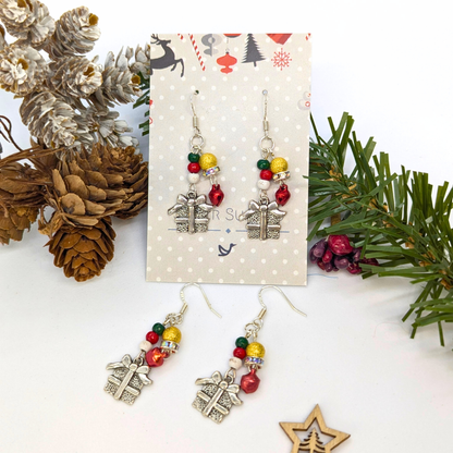 Gift Box Dangle Earrings by Silver Sunbird feature sterling silver hooks, silver present charms, and colorful beads. Displayed with pinecones and evergreens, each earring card is adorned with a snowflake pattern.