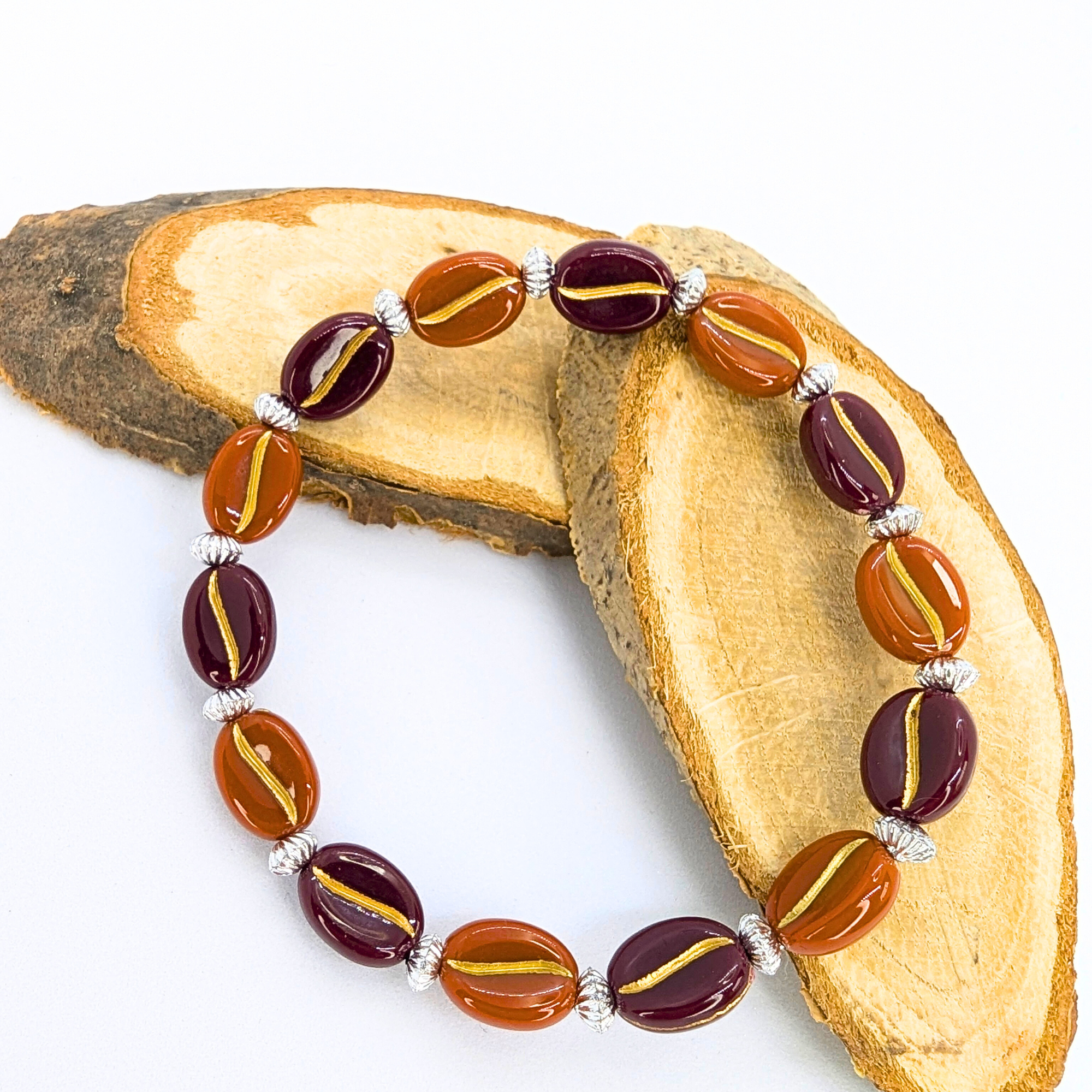 The Silver Sunbird Coffee Bean Bracelet showcases alternating brown and deep purple oval beads with gold accents, elegantly displayed on light wood against a white backdrop. Enhanced by silver spacer beads, it&