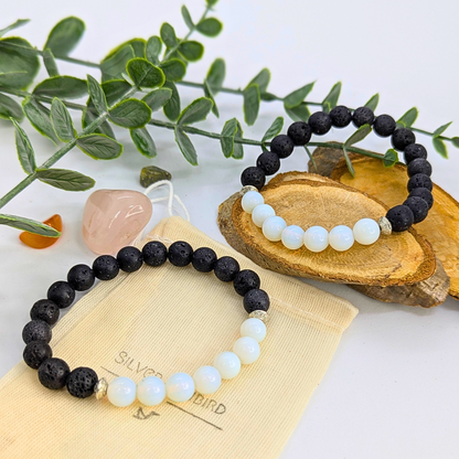 Two Silver Sunbird Opal and Lava Stone Bracelets rest on a wooden slab and pouch, with greenery and two crystals—one pink, one orange—in the bright, minimal background.