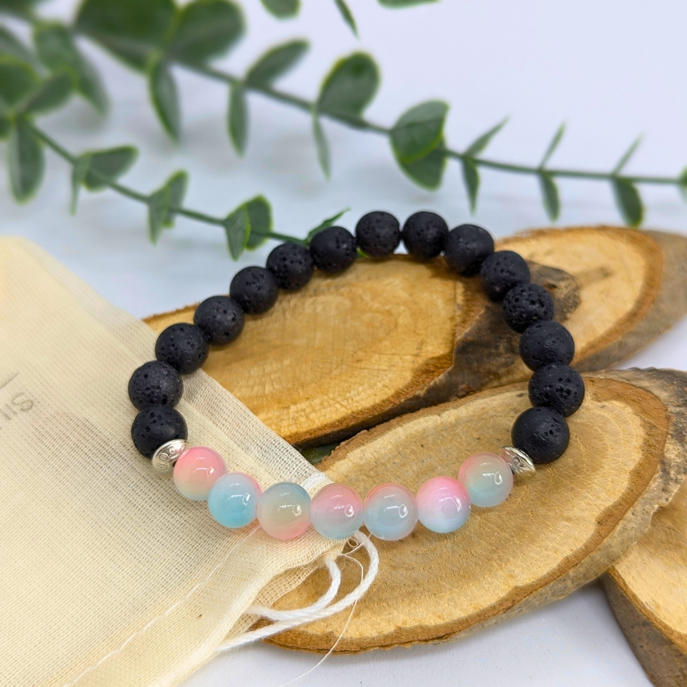 The Silver Sunbird Mermaid-Inspired Lava Stone Bracelet features black lava stones and pastel blue, pink, and white beads. Displayed on wooden slices with a cream drawstring pouch, it&