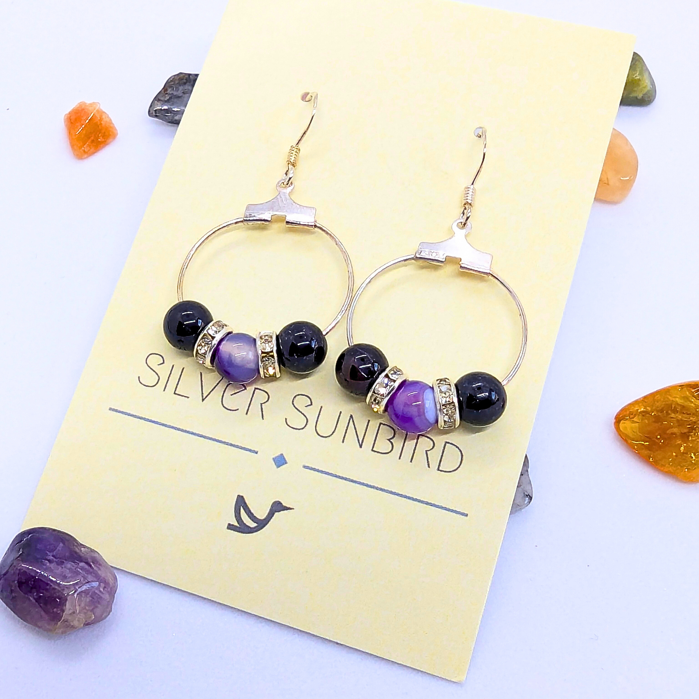 A pair of Amethyst Statement Large Earrings displayed on a yellow Silver Sunbird card. The earrings feature a circular design with three purple gemstone with silver rhinestone spacers. Nearby are scattered polished natural gemstones.