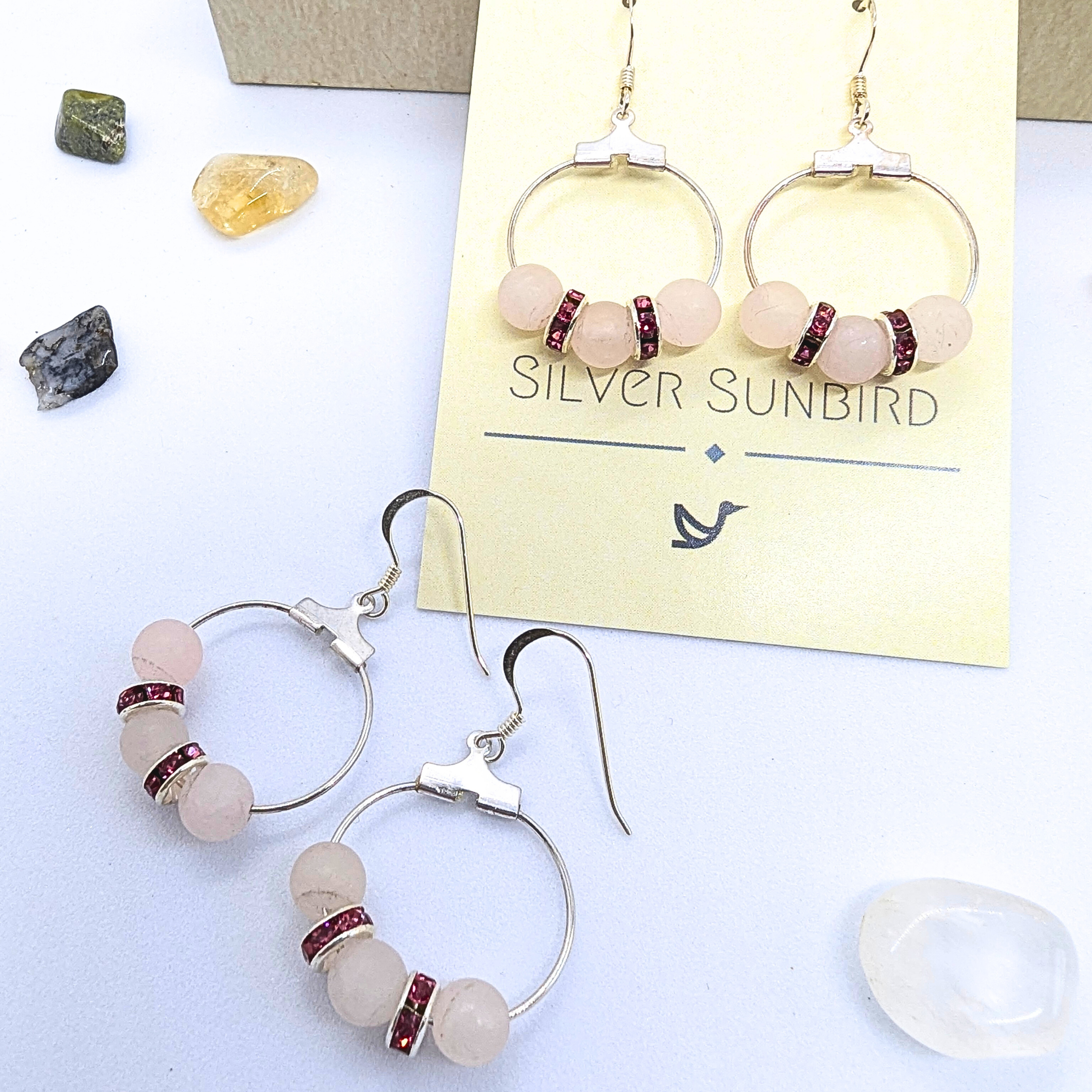 A pair of Rose Quartz Statement Earrings from Silver Sunbird, each adorned with three rose quartz gemstones and two dark red spacer beads. One pair hangs from a card, while the other lies flat. Small crystals in yellow, green, and white surround these hypoallergenic sterling silver earrings.