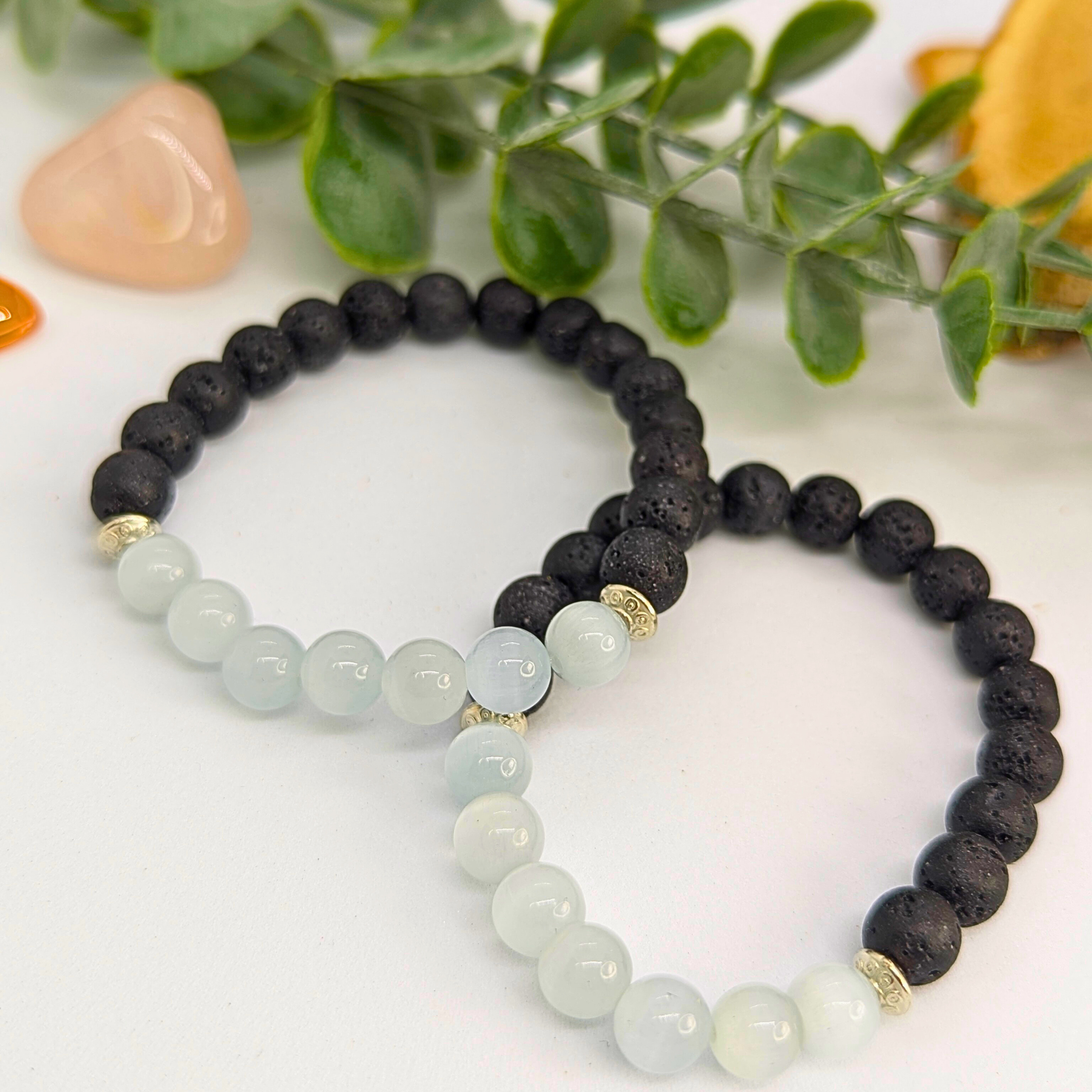 Two Moonstone and Lava Bracelets by Silver Sunbird are displayed on a white surface, each combining black lava stone beads with light blue moonstones. Green leaves and decorative stones provide a stylish background.